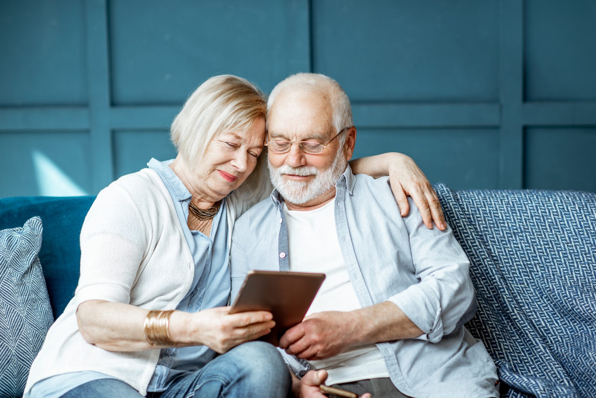 Elderly couple sitting together on a sofa, looking at a tablet with a content expression.