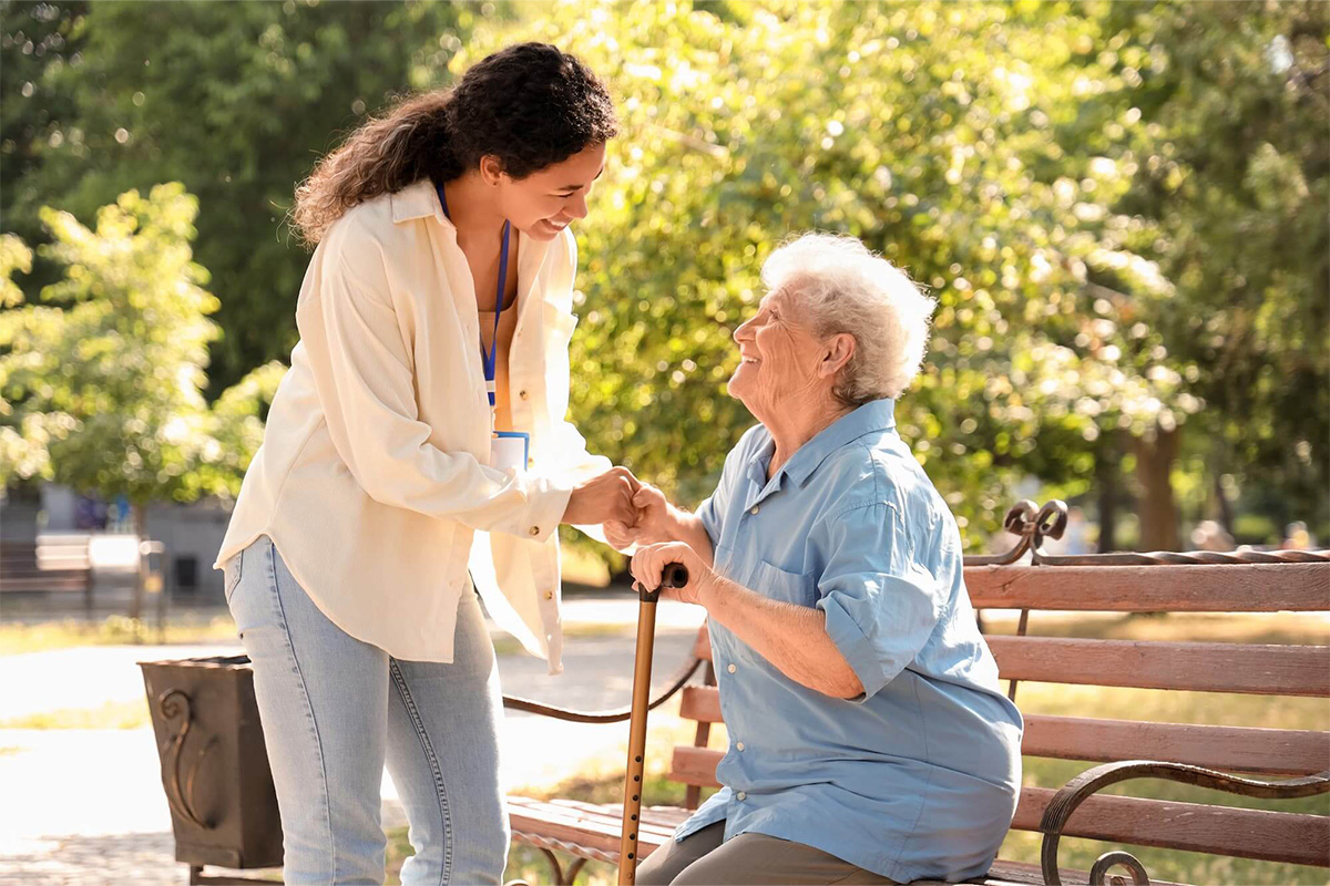 Senior woman sits on an outdoor bench smiling up at younger female caretaker.